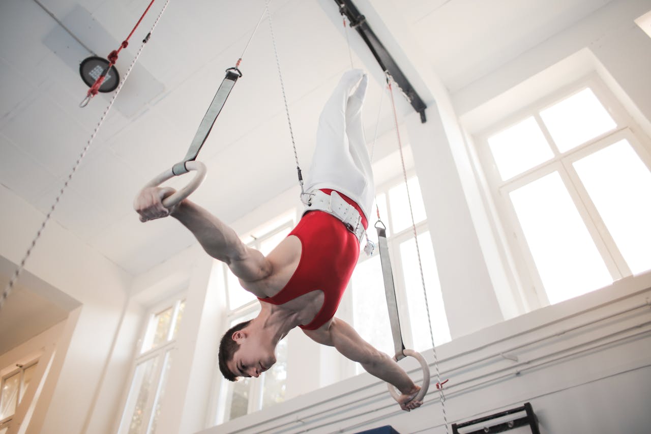 gallery-05 A male gymnast performs an upside-down pose on rings in a well-lit gym interior.