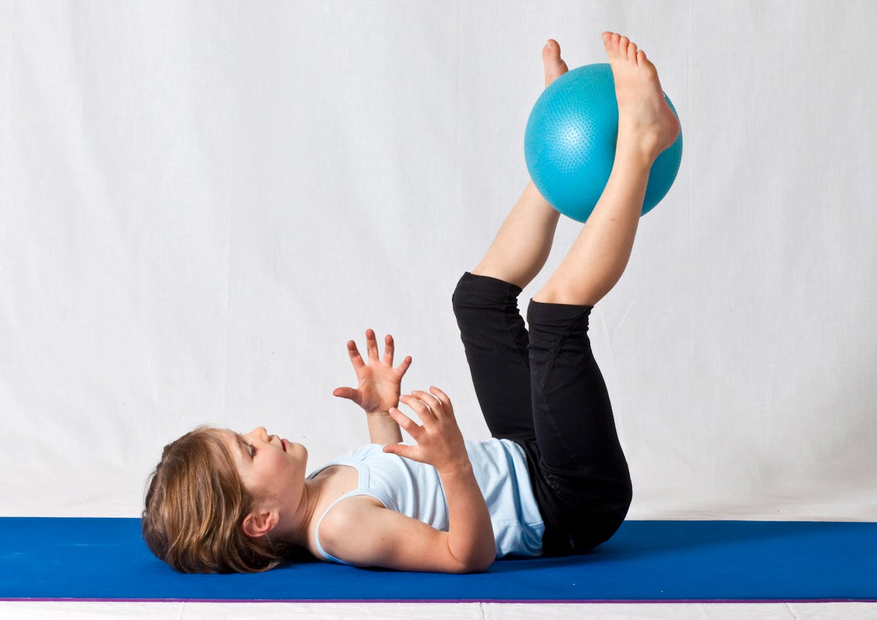 our-services-01 Young girl practicing balance exercises with a blue yoga ball indoors on a mat.