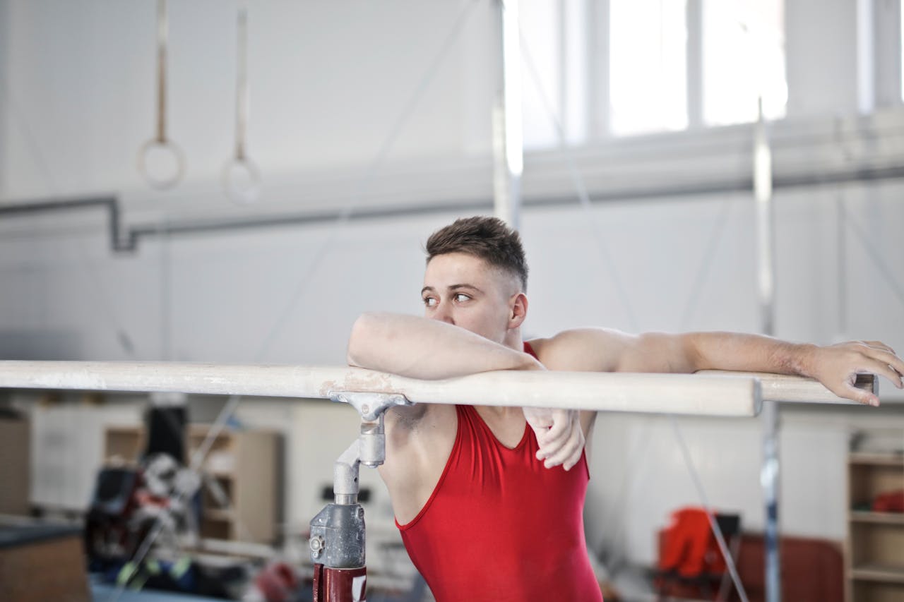 Male gymnast in red attire resting on parallel bars in a gym, showing strength and focus.