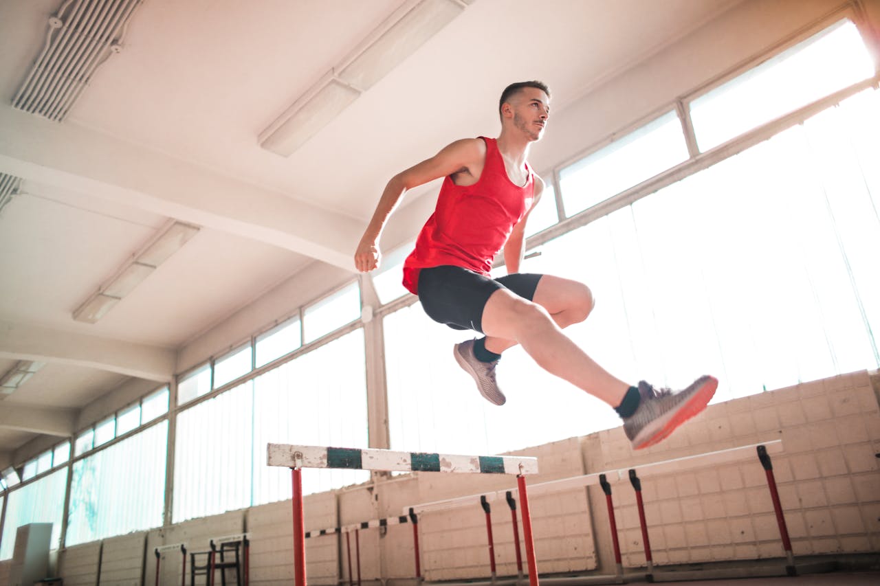 gallery-04 A young male athlete in a red outfit jumps over a hurdle indoors, showcasing agility and determination.