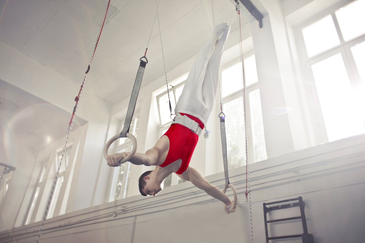 about-us-02 A male gymnast exercises on rings in an indoor gym, showcasing strength and flexibility.